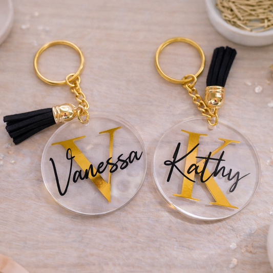 Two keychains with 'Vanessa' and 'Kathy' on clear discs with gold lettering, black tassels, on a decorative background.