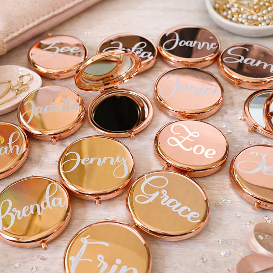 Compact mirrors with names engraved on their lids arranged on a surface.