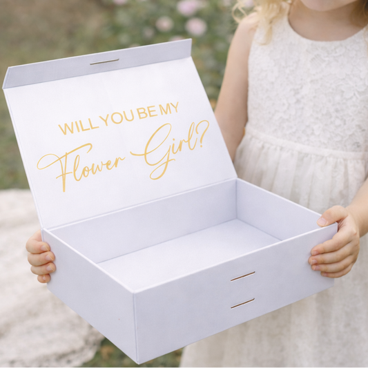 White box with 'Will you be my Flower Girl?' text held by a young girl in a white dress outdoors.