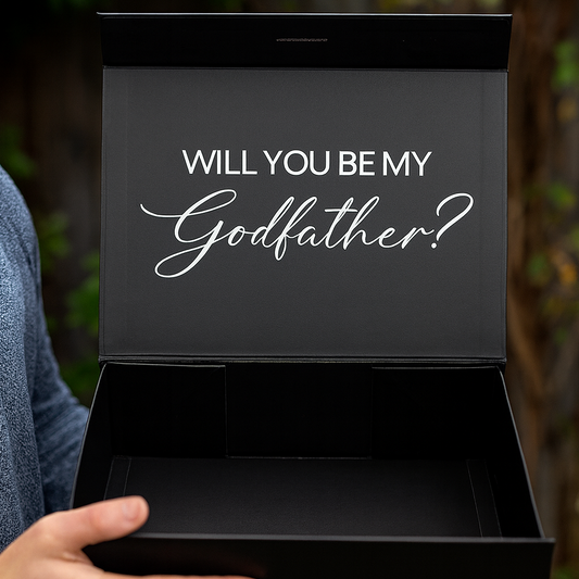 Person holding a black box with 'Will You Be My Godfather?' text against a blurred natural background