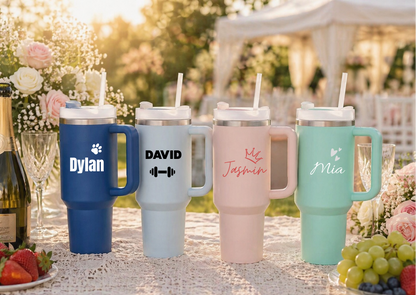 Four insulated travel mugs with personalized names on a table outdoors.