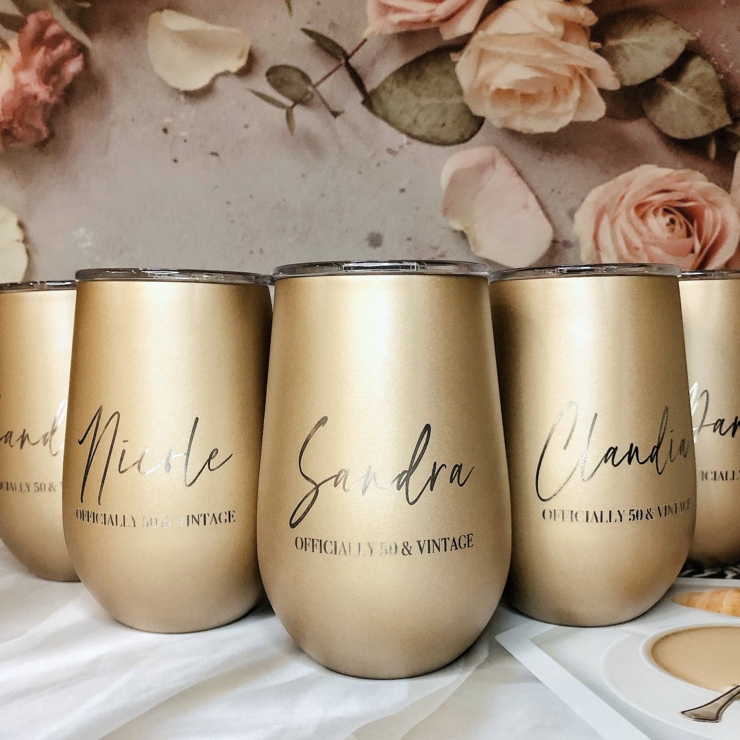 A set of four engraved gold stainless steel travel tumblers with names on them, displayed on a table with a floral background and a small amount of liquid in them.