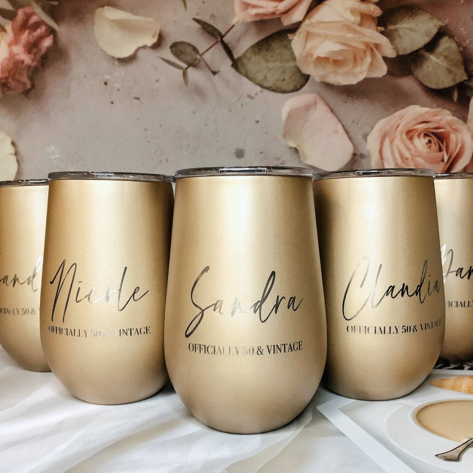 A set of four engraved gold stainless steel travel tumblers with names on them, displayed on a table with a floral background and a small amount of liquid in them.