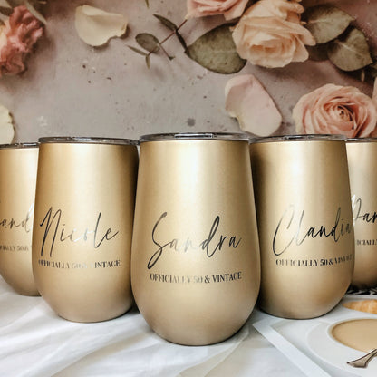 A set of four engraved gold stainless steel travel tumblers with names on them, displayed on a table with a floral background and a small amount of liquid in them.