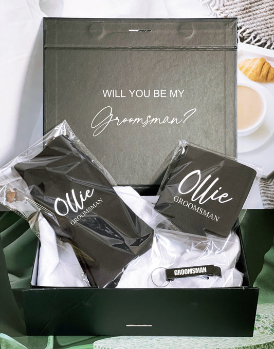 A gift box with the text 'WILL YOU BE MY GROOMSMAN?' on the lid, containing personalized socks, a stubby holder, and a bottle opener, all customized with the name 'Ollie'.