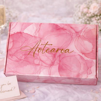 Pink floral box with 'Aotearoa' text, champagne flute, ring, and card on a decorative background