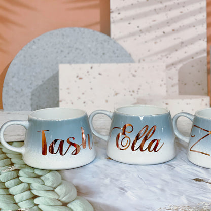 Three stoneware mugs with hand-glazed finish, personalized with vinyl names 'Tash', 'Shella', and 'Zella' in a blue and cream color scheme, displayed on a table with a decorative palm leaf in the background.