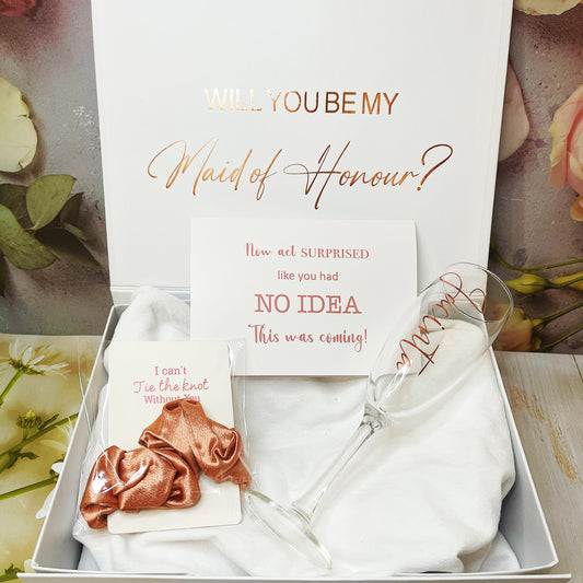 A gift box proposal set for bridesmaids including a personalized box with text, a champagne flute, a silk ribbon, and a card with the question 'Will you be my Maid of Honour?'.