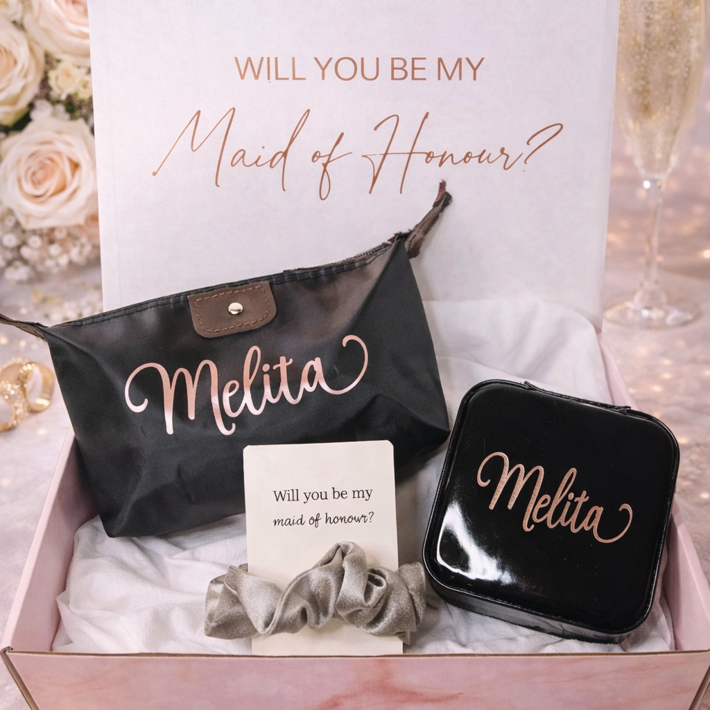 Gift set with 'Will you be my Maid of Honour?' card, black pouch, and jewelry box, surrounded by flowers and champagne glasses.