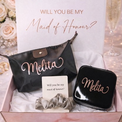 Gift set with 'Will you be my Maid of Honour?' card, black pouch, and jewelry box, surrounded by flowers and champagne glasses.