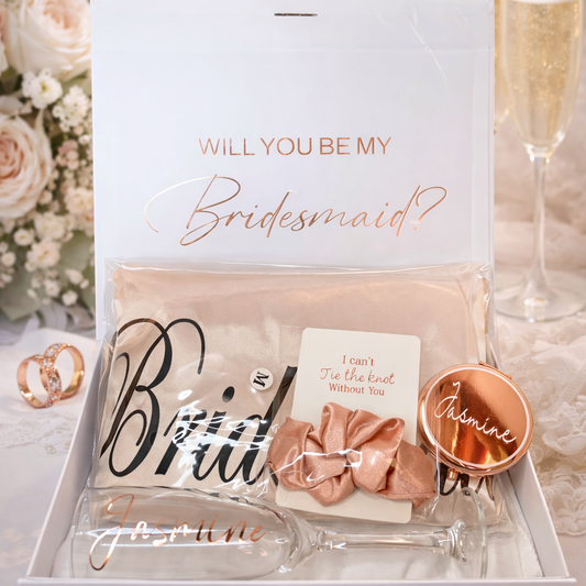 Gift box with 'Will you be my bridesmaid?' card and decorative items on a table with flowers and champagne.