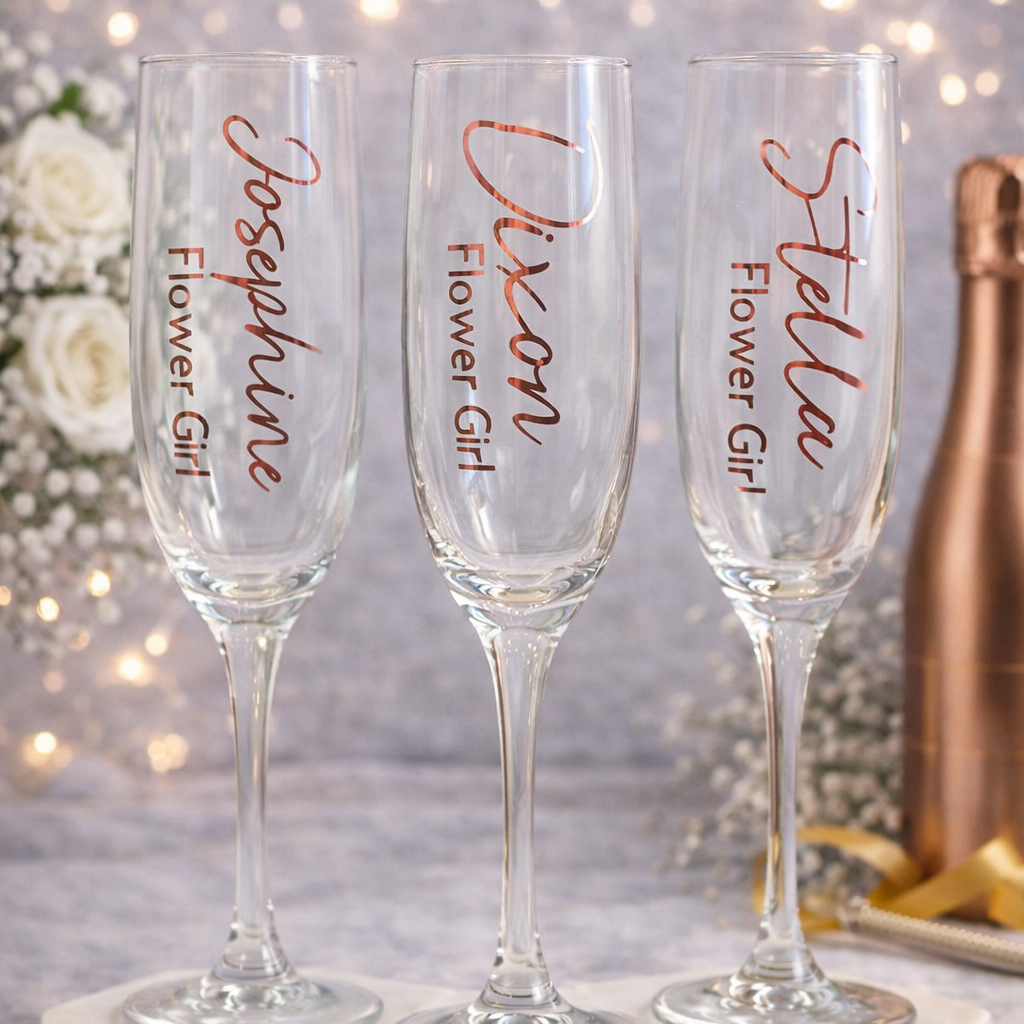 Three champagne flutes with personalized names on a marble stand against a blurred decorative background.