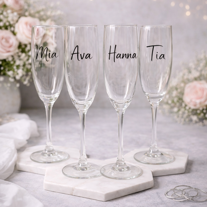 Four champagne flutes with names 'Mia', 'Ava', 'Hanna', and 'Tia' on a marble surface with flowers and lights in the background.