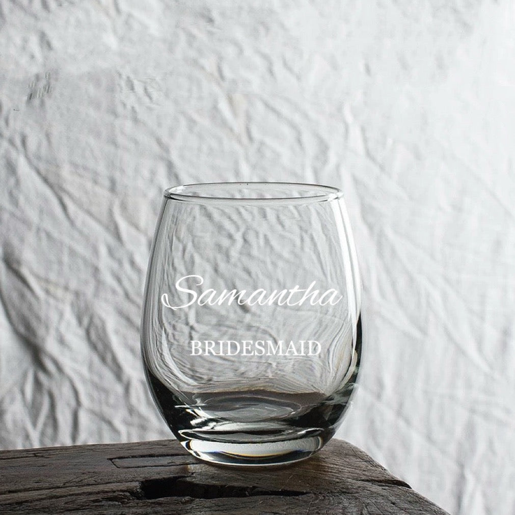 A clear stemless wine glass with the engraved text 'Samantha' and 'BRIDESMAID' on it, placed on a wooden surface with a textured background.