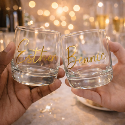 Two hands holding personalized glass tumblers with 'Esther' and 'Bennie' on a blurred background.