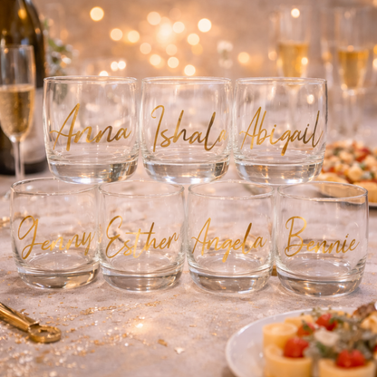 Clear glasses with gold names on a festive table with blurred lights and food.