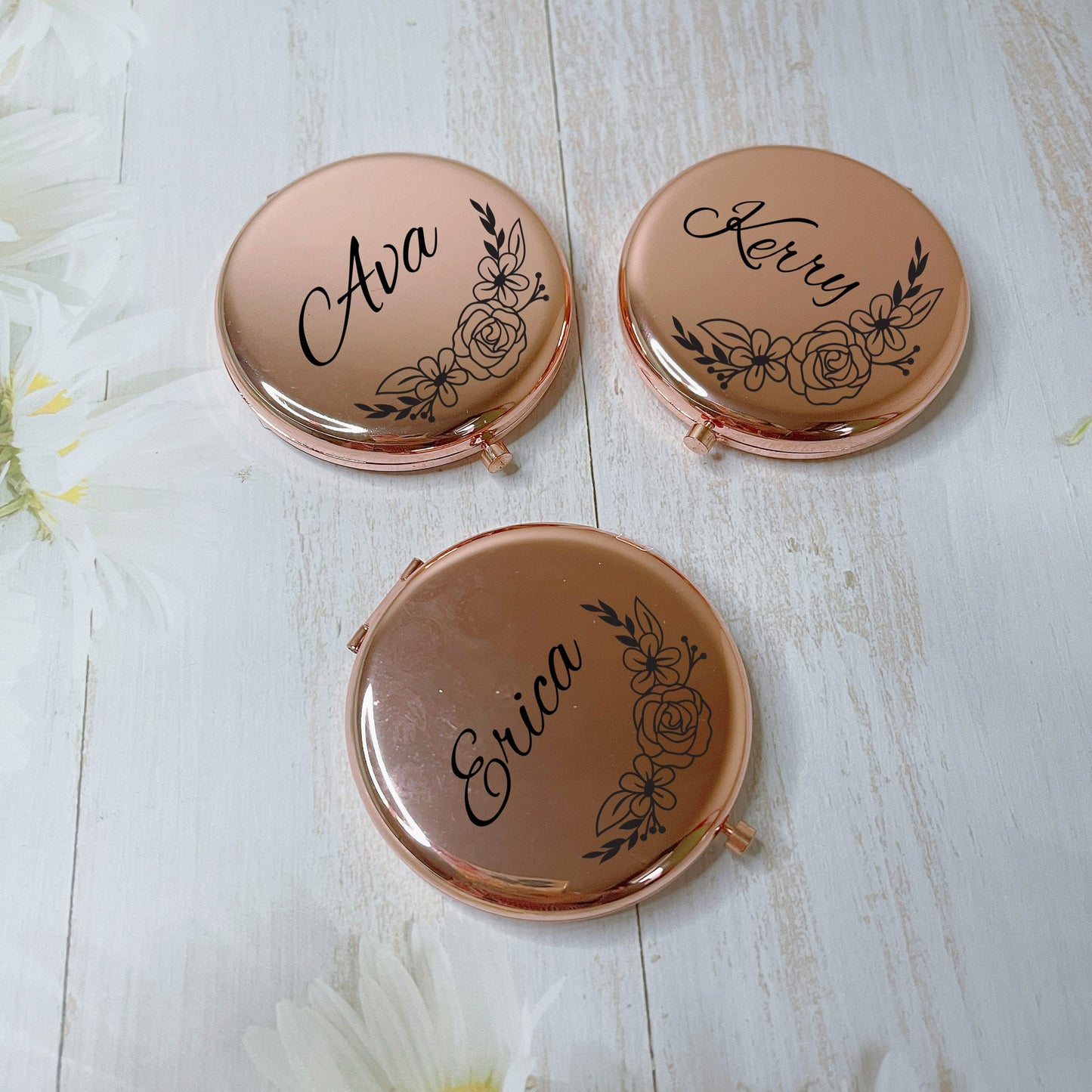 Three rose gold compact mirrors with names engraved on them, placed on a white wooden surface with flowers around.