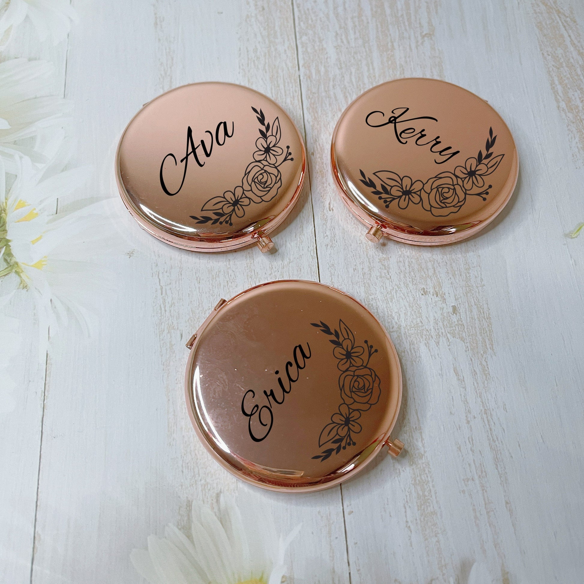Three rose gold compact mirrors with names engraved on them, placed on a white wooden surface with flowers around.