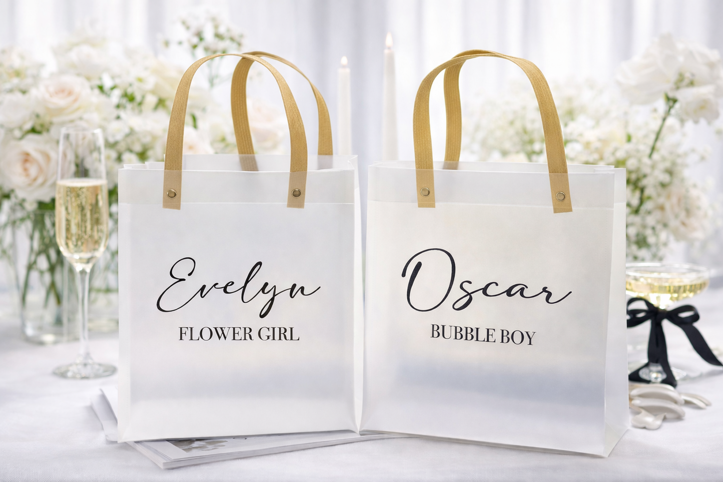 Two white bags with gold handles, one labeled 'Evelyn Flower Girl' and the other 'Oscar Bubble Boy', on a table with flowers and champagne.