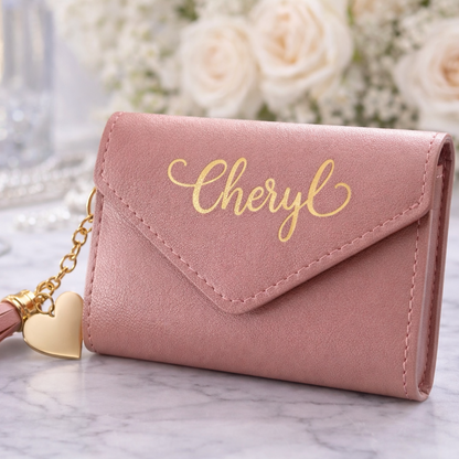 Pink wallet with 'Cheryl' engraving and tassel on a marble surface with flowers and glasses in the background