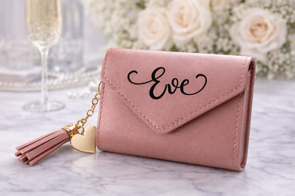 Pink wallet with 'Eve' engraving and tassel on a marble surface with flowers and glasses in the background