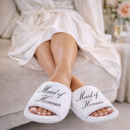 White slippers with 'Maid of Honour' text worn by a person in a white robe.
