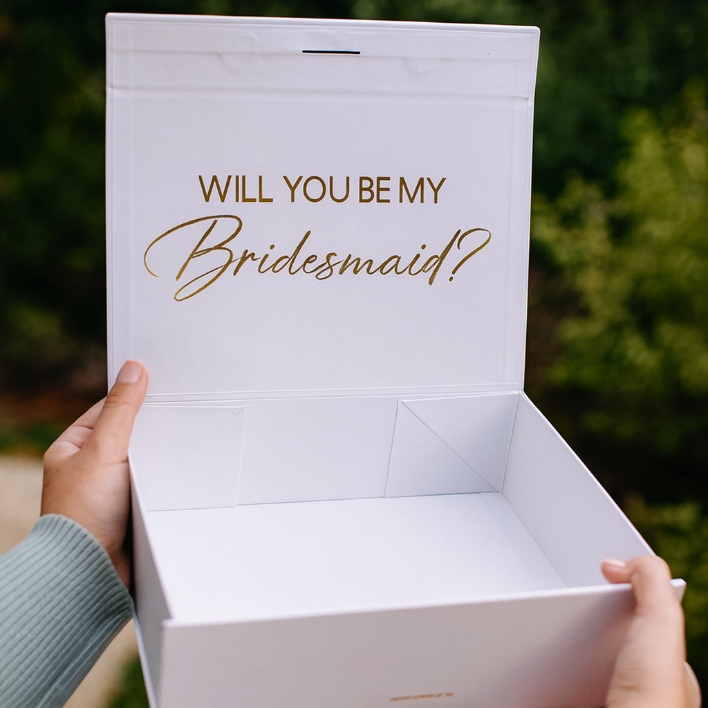 White box with 'Will You Be My Bridesmaid?' text held by two hands against a blurred green background.