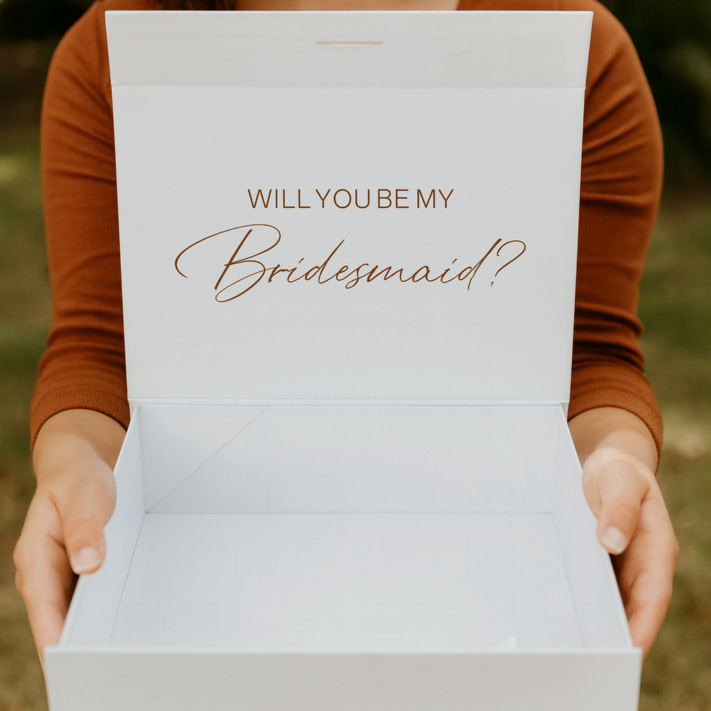 Person holding a box with 'WILL YOU BE MY Bridesmaid?' text outdoors.