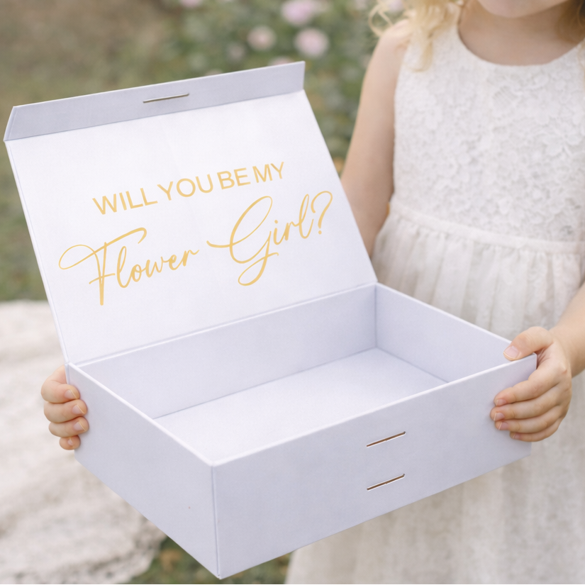 White box with 'Will you be my Flower Girl?' text held by a young girl in a white dress outdoors.