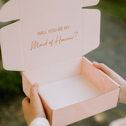 Pink box with 'Will you be my Maid of Honor?' text held by a person.