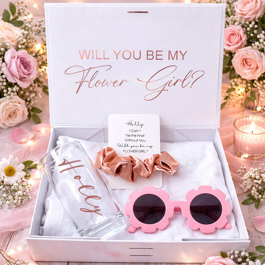 Gift set with pink sunglasses, hair accessories, and card in a box with 'Will You Be My Flower Girl?' text, surrounded by flowers and lights.