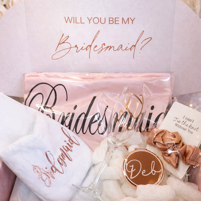 Gift box with 'Will you be my bridesmaid?' and 'Bridesmaid' text, flowers, and a card on a soft background.