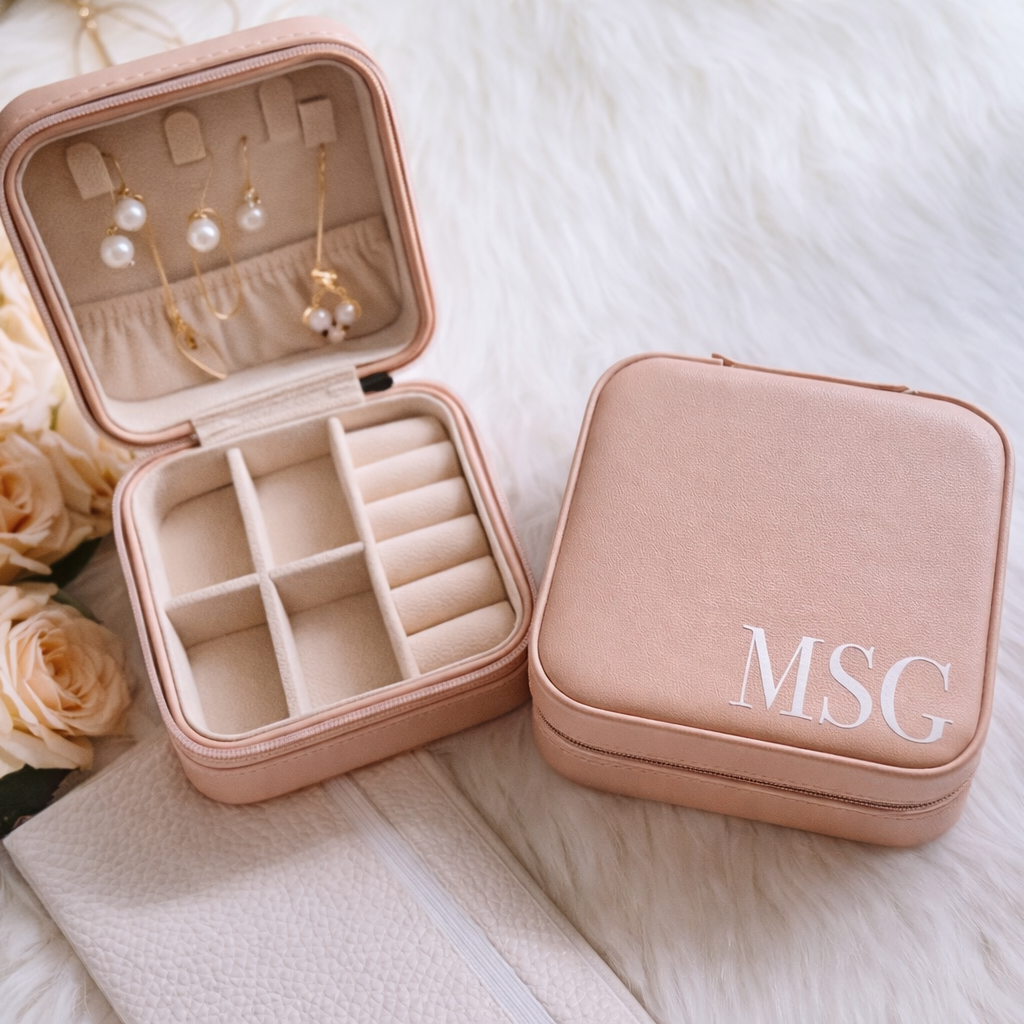 Pink jewelry case with 'MSG' monogram, open to display earrings and rings, on a white surface with flowers.