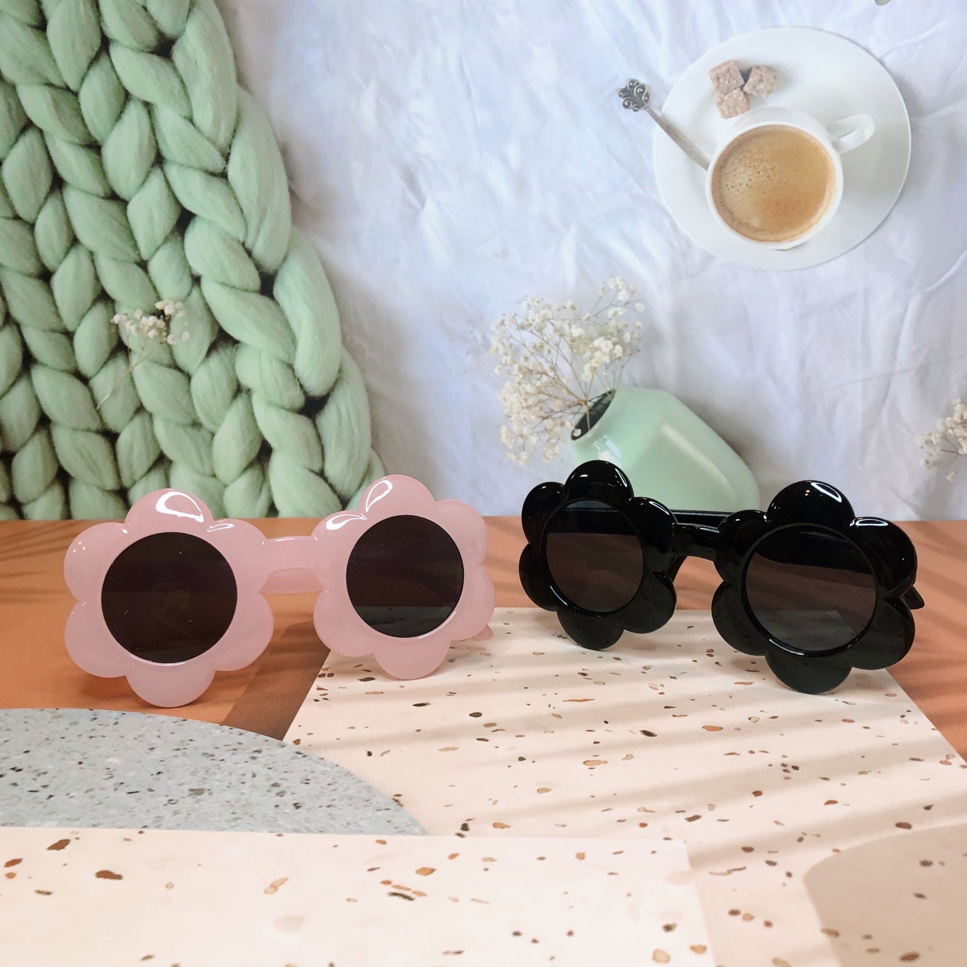 Two pairs of heart-shaped sunglasses for kids, one in pink and one in black, displayed on a surface with a cup of tea and a knitted blanket in the background.