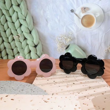 Two pairs of heart-shaped sunglasses for kids, one in pink and one in black, displayed on a surface with a cup of tea and a knitted blanket in the background.