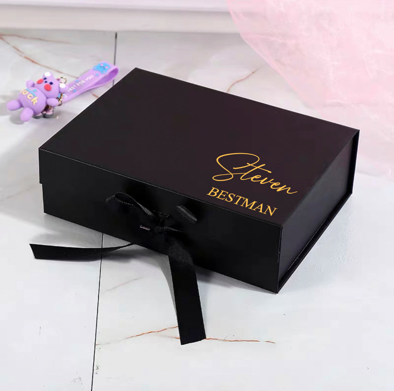 A black gift box with a personalized golden label that reads 'Steven Bestman', adorned with a subtle black ribbon bow.