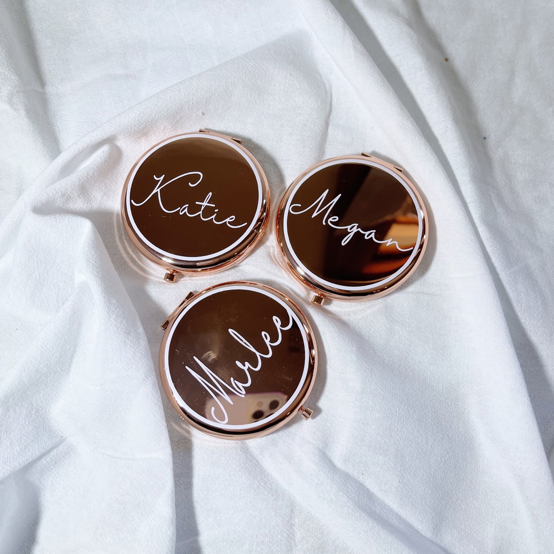 Three personalized rose gold mirrors with the names 'Kate', 'Megan', and 'Mia' written on them, placed on a white fabric surface.