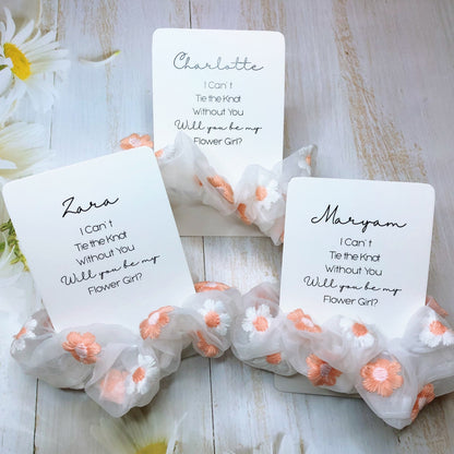A set of satin scrunchies with personalized gift cards tied to them, presented on a wooden surface with flowers around.