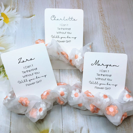 A set of satin scrunchies with personalized gift cards tied to them, presented on a wooden surface with flowers around.
