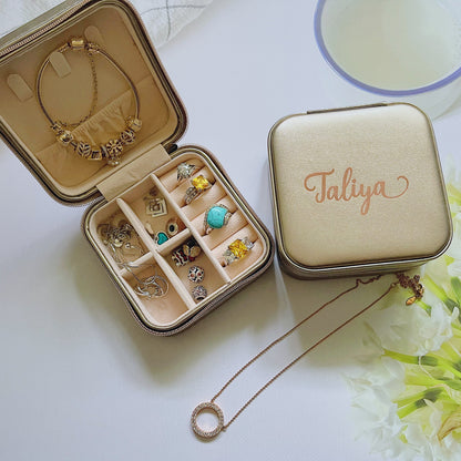 A compact travel jewellery box opened to display multiple compartments with jewelry items like rings and earrings. The box is beige with a personalized name 'Taliya' written in a stylish font on the lid.
